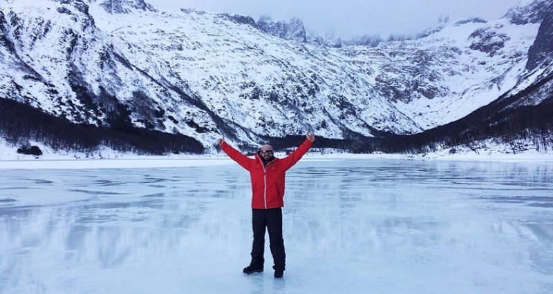 Inverno em Ushuaia - Turista fazendo pose para a foto no centro da imagem em cima do lago congelado. Ao fundo montanhas cobertas de neve.