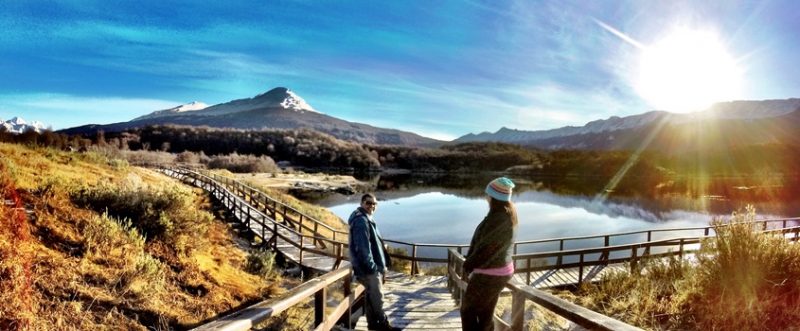 Ushuaia Verão - Casal em cima da passarela olhando um para o outro. Ao fundo uma lagoa e atras do rio montanhas. Céu azul com algumas nuvens na cor roxa e o Sol saindo por trás das montanhas.