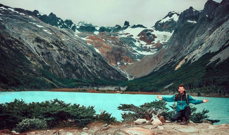 Homem posando a frente do Lago Esmeralda em ushuaia no verão. Ao fundo, montanhas cobertas de neve completam a vista.